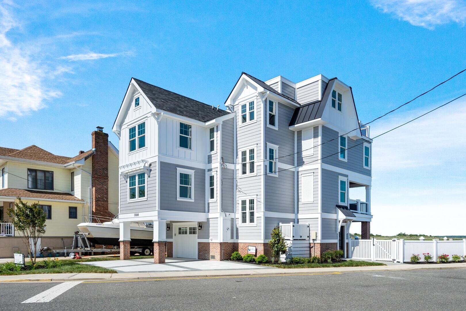A striking contemporary four-story vacation rental at 5800 Lake Road in Wildwood Crest, featuring a sleek grey and white exterior with a boat parked underneath. This luxury waterfront property offers private pool access and stunning bay views for a premier Long & Foster Vacation Rentals experience.