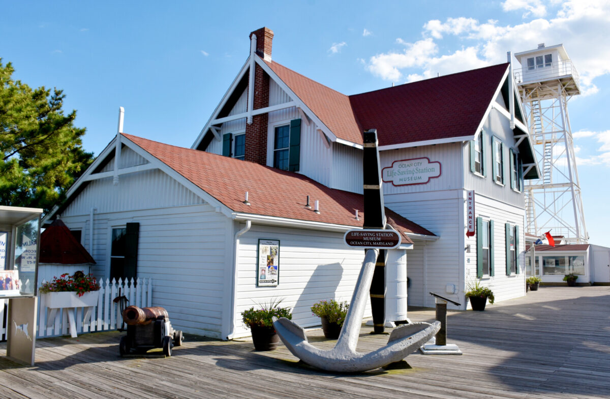 The historic white and red Ocean City Life-Saving Station Museum located on the boardwalk, featuring a large anchor and antique cannon out front. This cultural landmark offers a glimpse into local maritime history for guests staying at a Long & Foster Vacation Rentals property in Ocean City, Maryland.