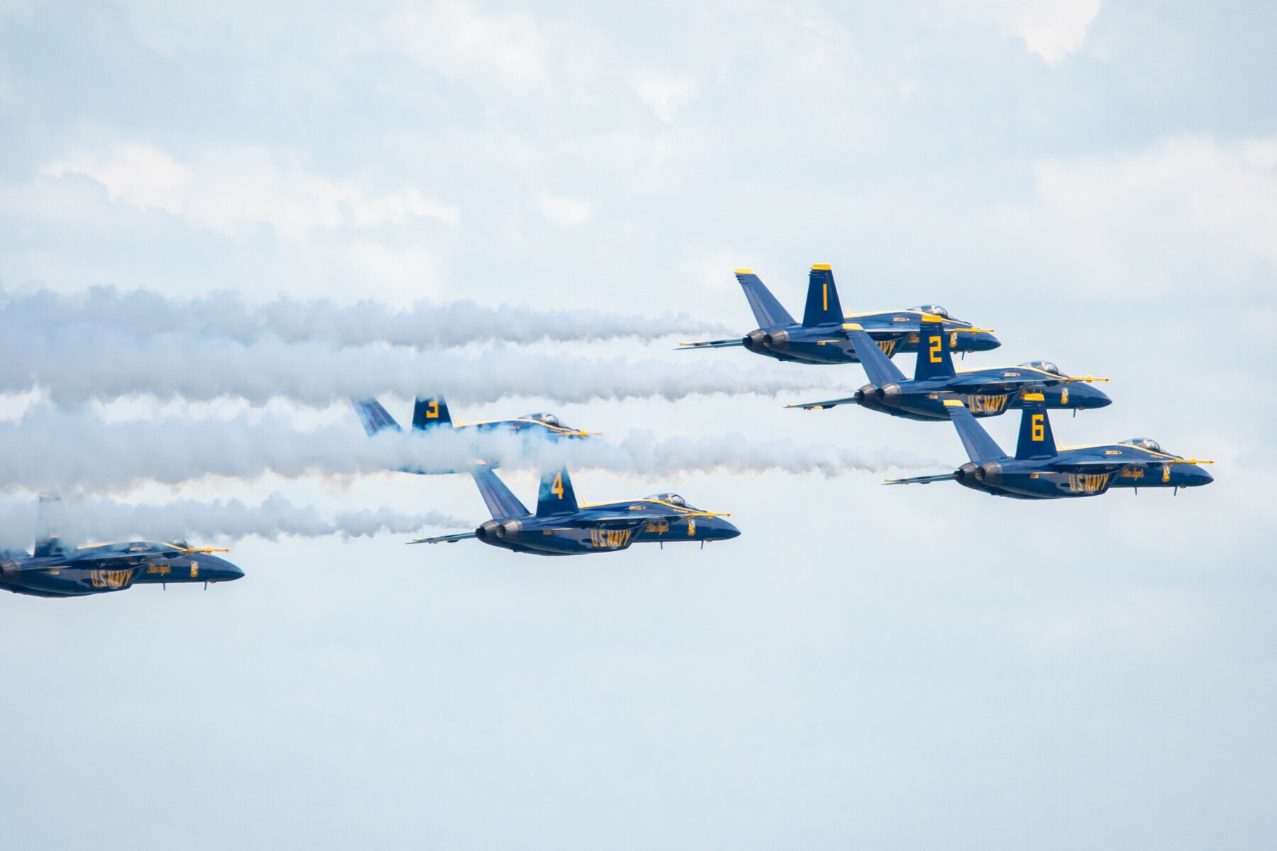 Blue Angels aircrafts in tight formation with white smoke trails across a blue sky.
